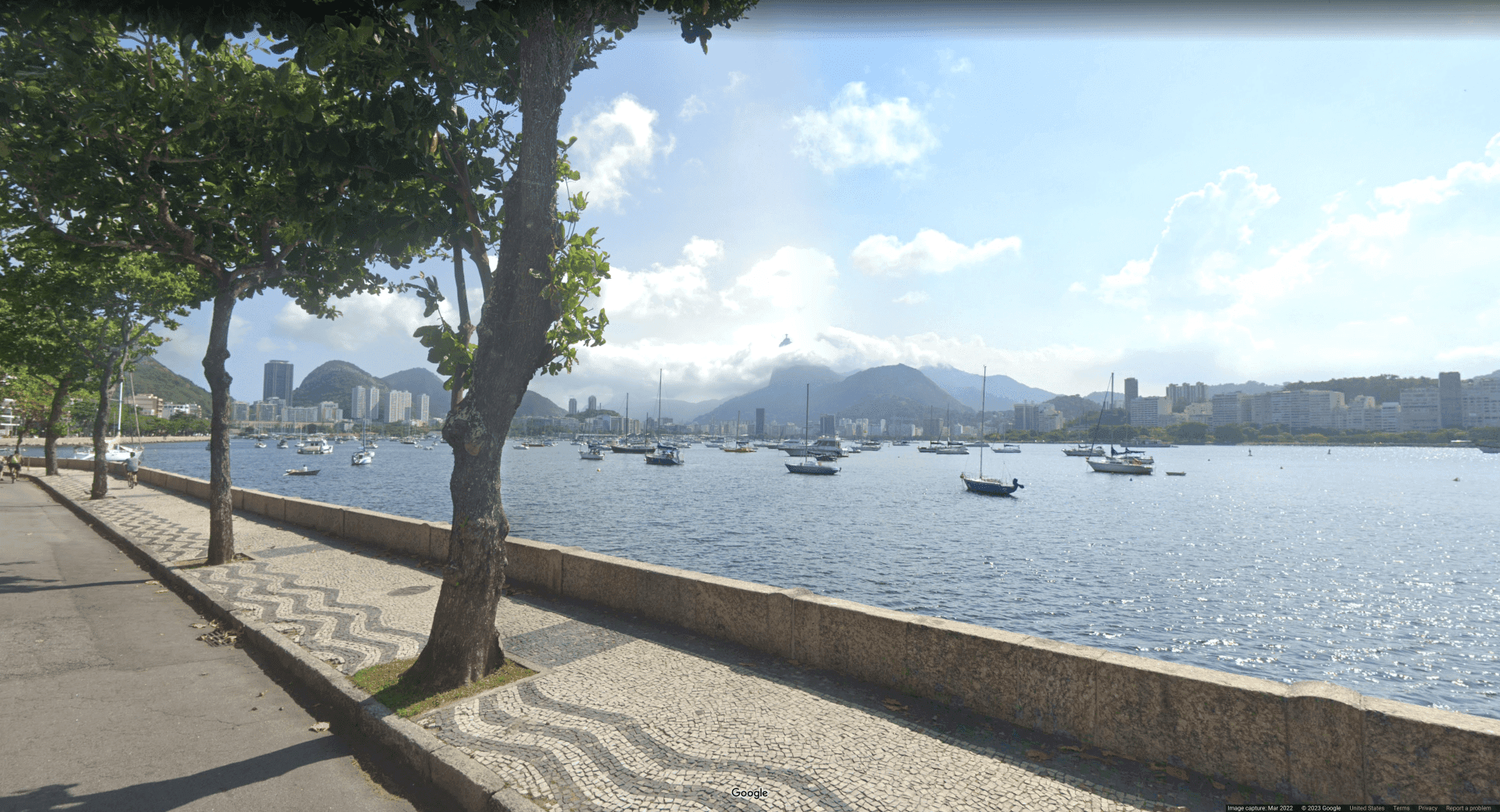 Picture of a waterfront walkway near a sea, with boats in a local harbor with overcast skies. A small statue is visible in the background, peeking above the clouds