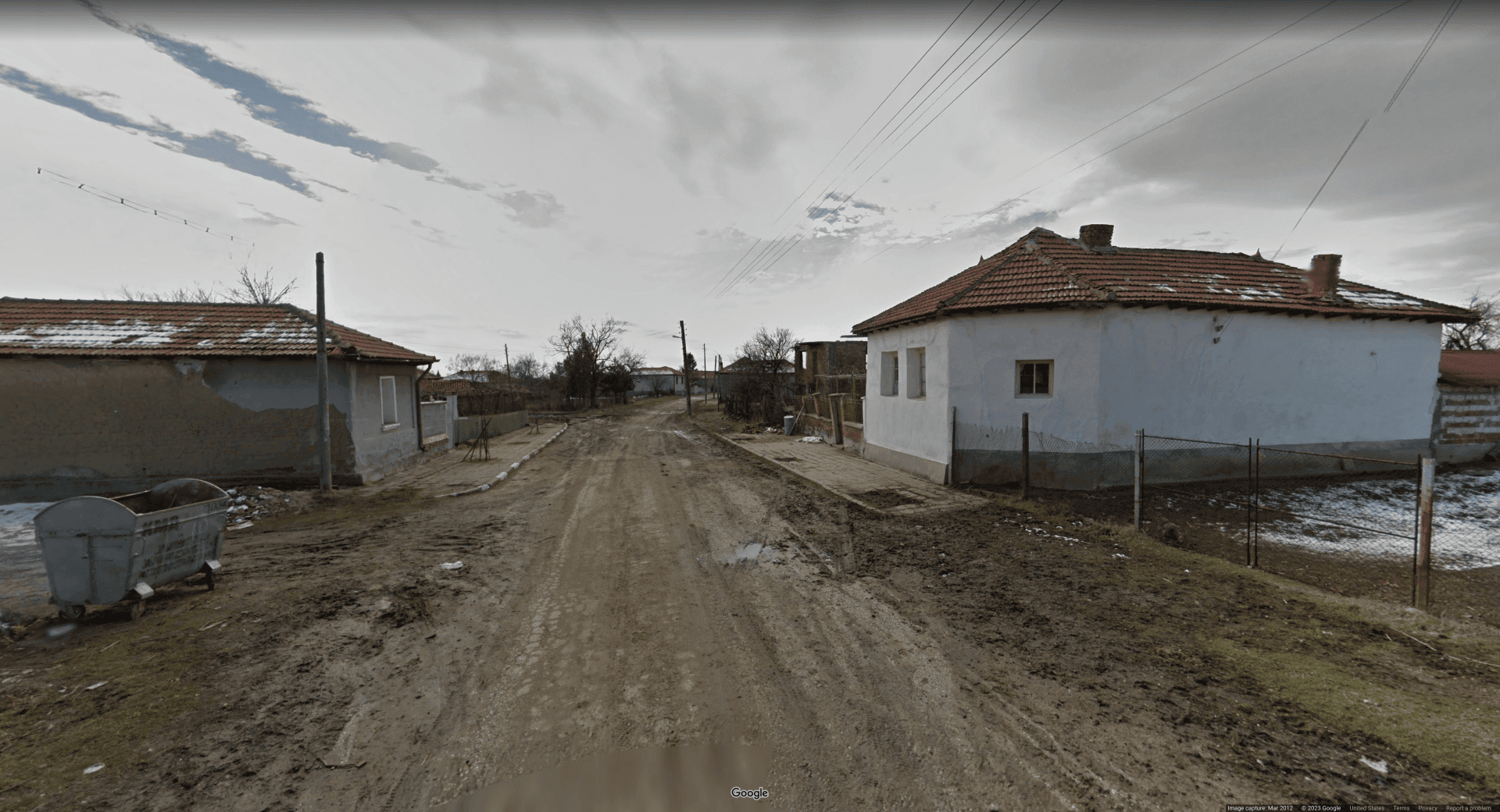 Picture of a dilapidated, bleak snowy area with a dirt road weaving through two red-tiled houses. A small metal dumpster rests on the left